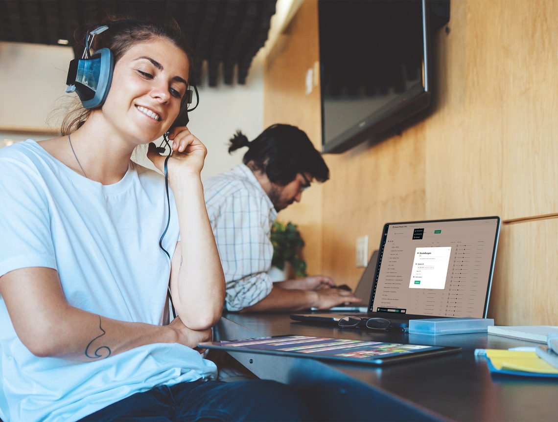 Eine junge, lächenlde Frau mit Headphones sitzt im Büro am Laptop und Tablet und hört einen Audiostream