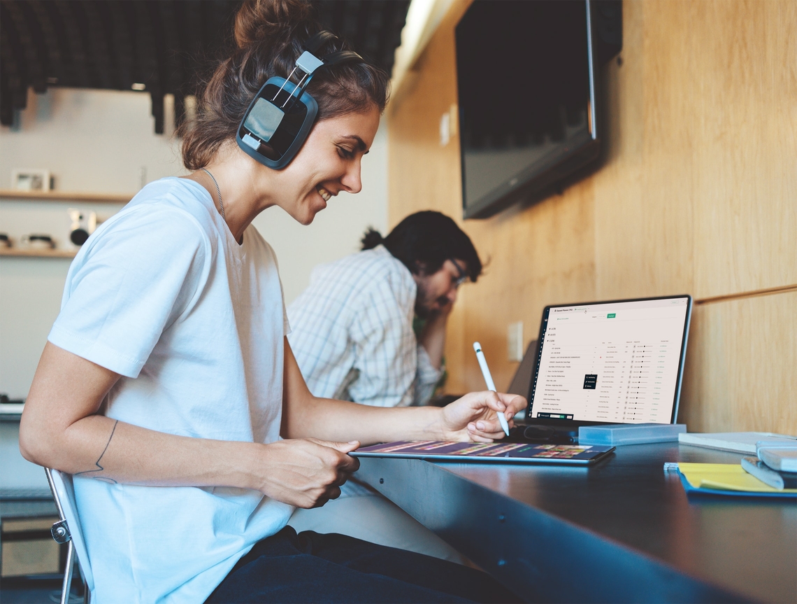 Eine junge, lächenlde Frau mit Headphones sitzt im Büro am Laptop und Tablet und hört einen Audiostream